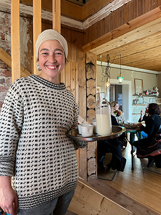 En glad kvinna med kaffebricka i handen hälsar välkommen till ett kafé.